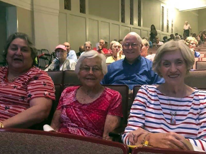 Four people sitting in theater seats, smiling. Others in the background.