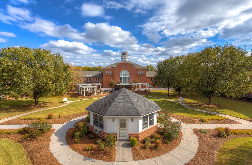 A large red brick building with a white gazebo in front, set in a landscaped yard under a blue sky.