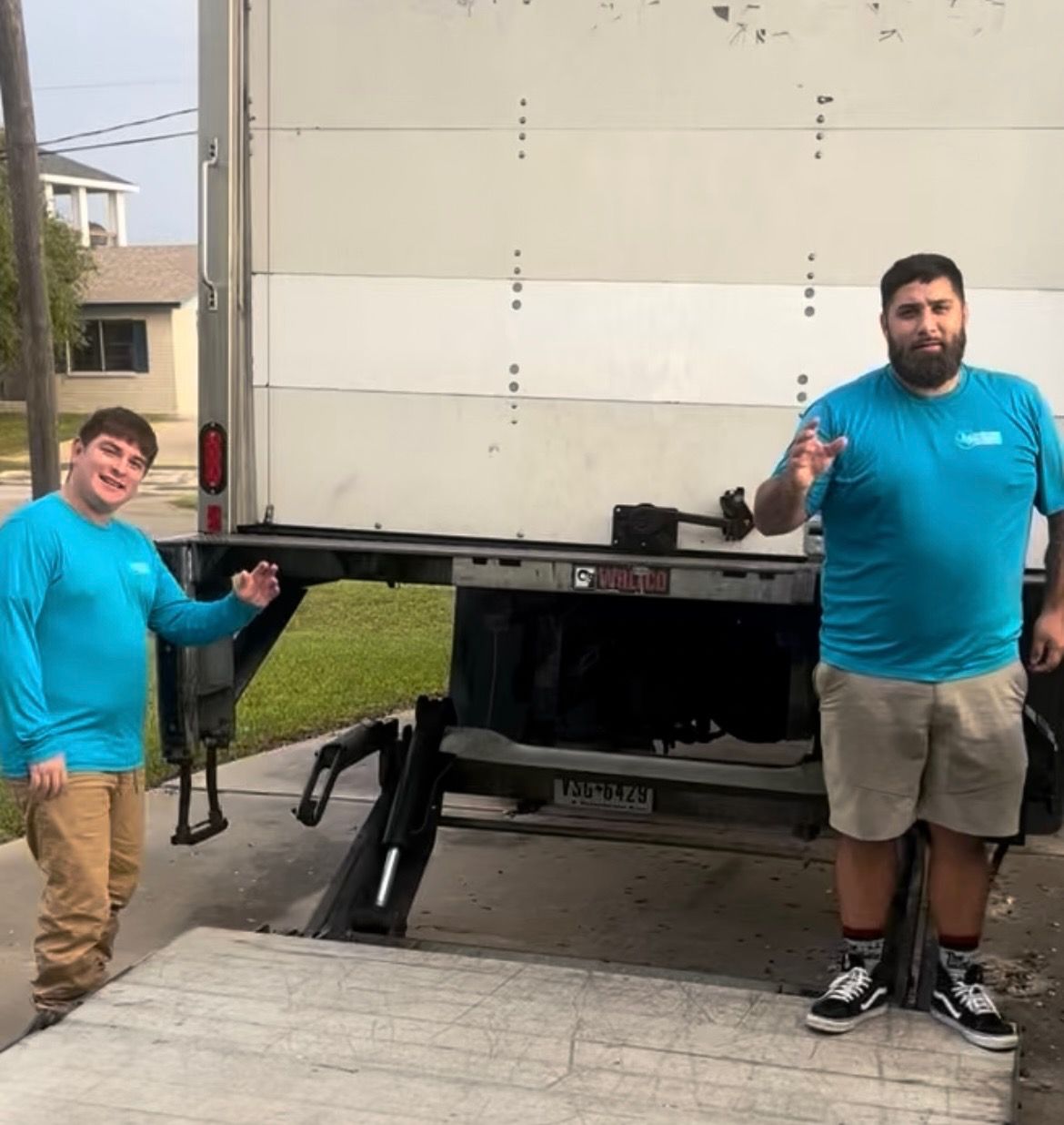 Two men in blue shirts and tan shorts stand next to an open moving truck, smiling and gesturing.
