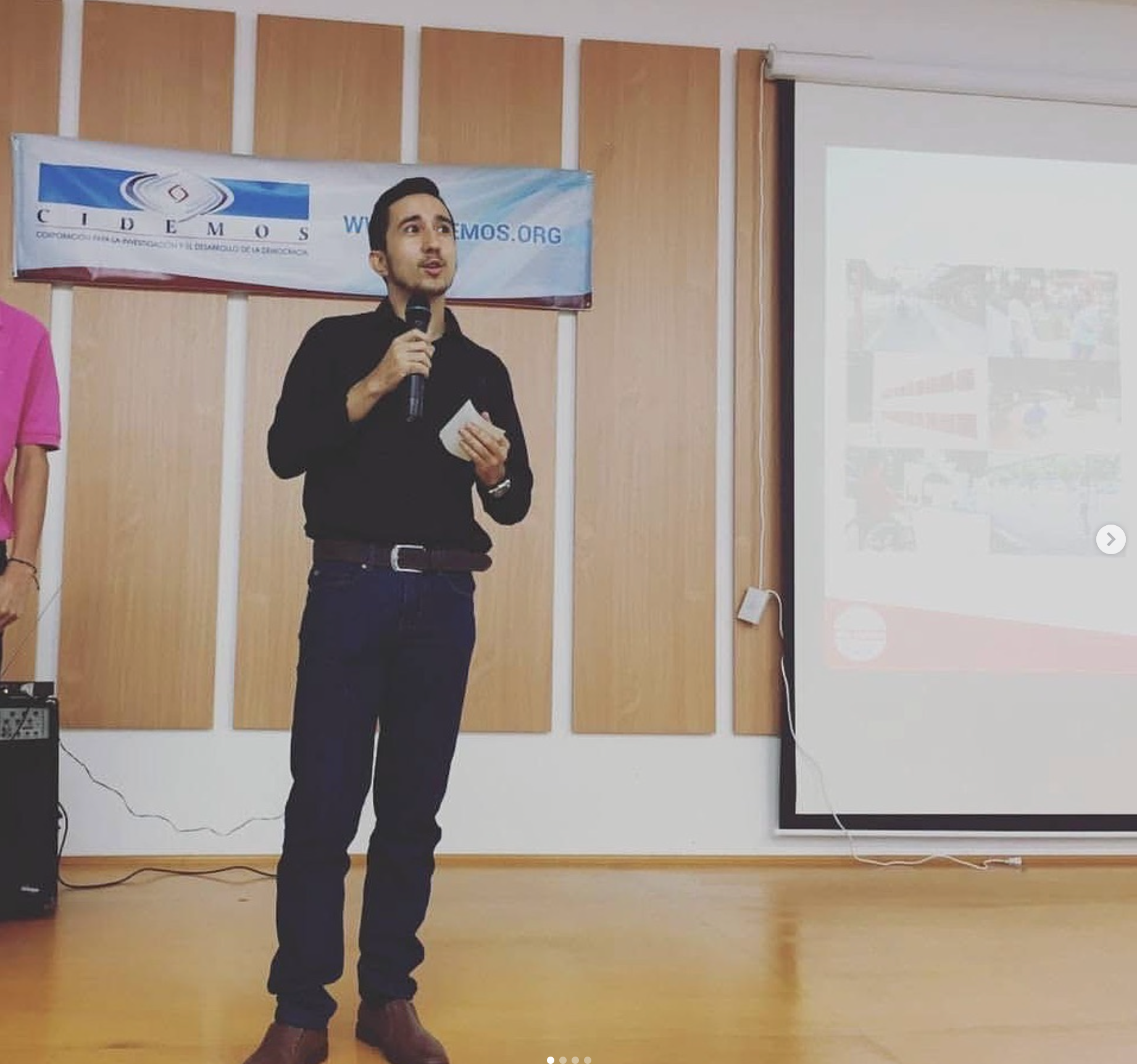 Man in black shirt speaks into microphone at a presentation; banner and screen in the background.