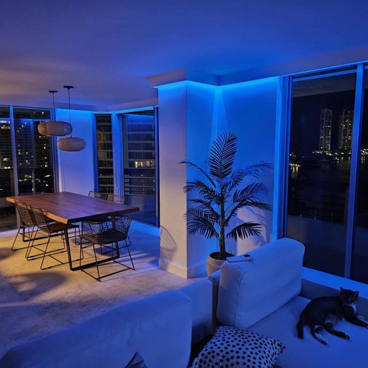 Living room at night with blue lighting, wooden table, city view, and cat on white couch.