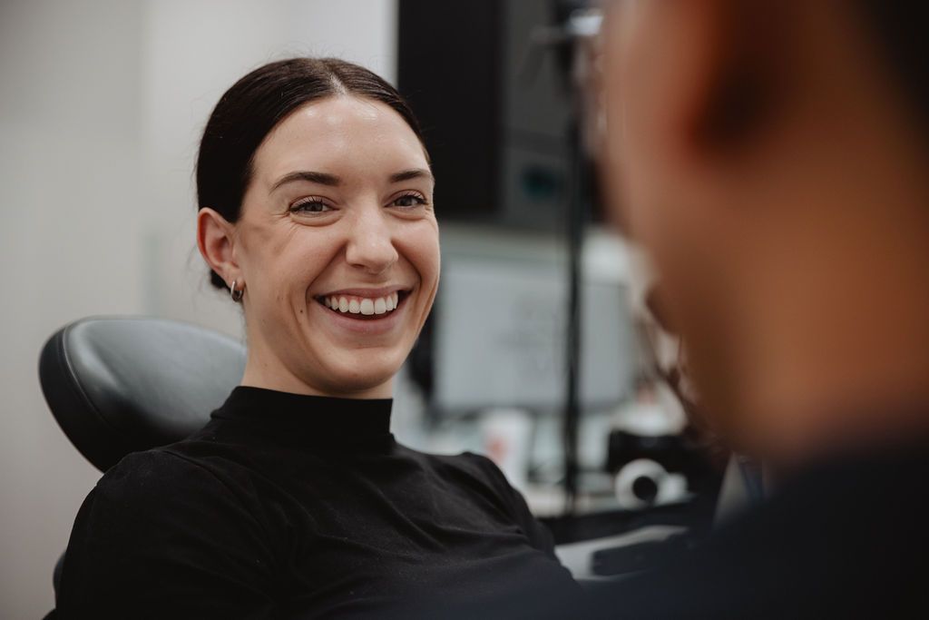 Woman smiling widely in a dental chair, facing a person out of frame, in a brightly lit office.