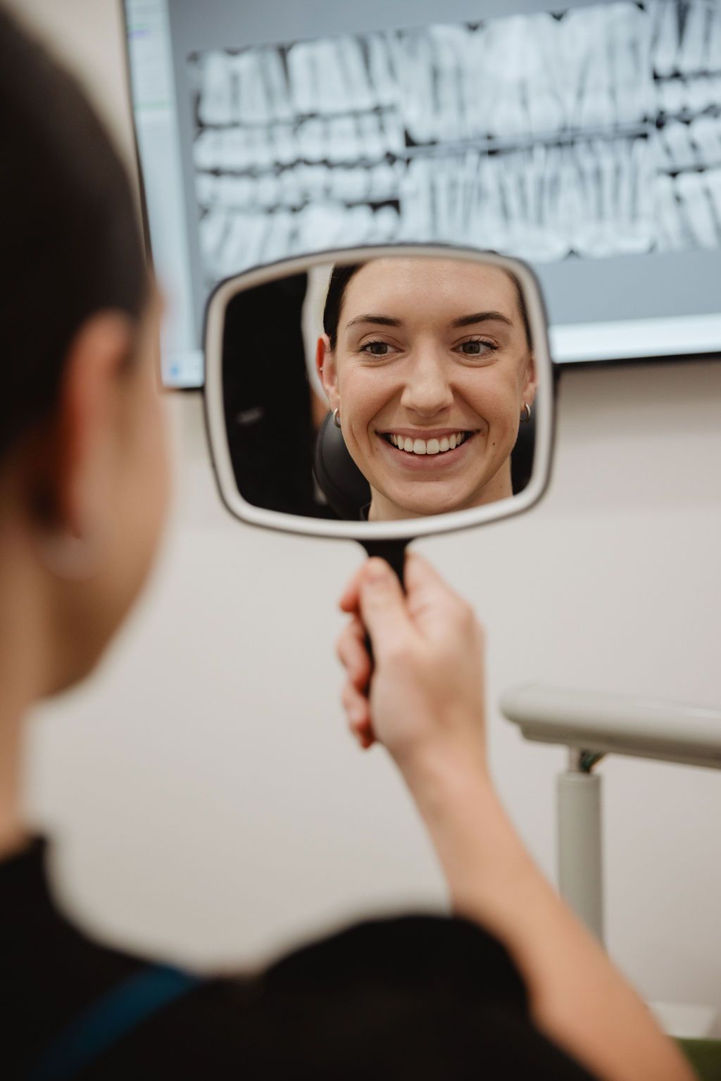 Woman smiling in a hand mirror, dental X-ray in background. Dentist's office setting.