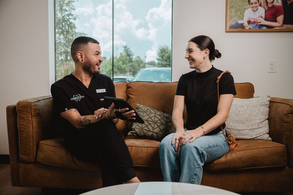 A person and medical professional converse on a brown couch. The medical professional holds a tablet and gestures.