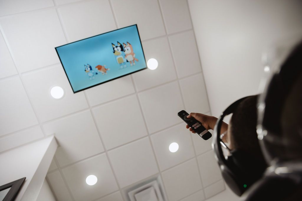 Person with headphones, pointing a remote at a ceiling-mounted TV playing Bluey cartoon. White tiled ceiling with lights.