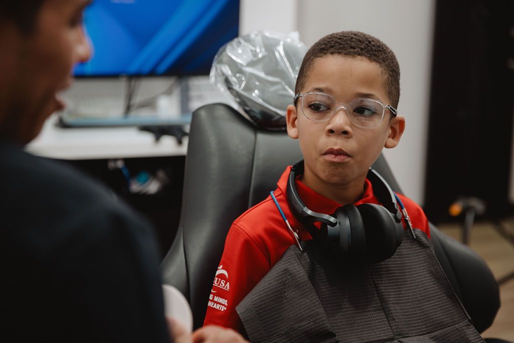 Boy in glasses listens to a person off-screen. Headphones around neck, red shirt, seated in chair.