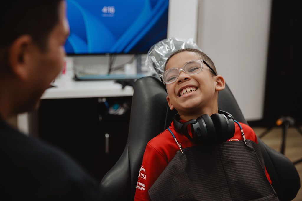 Boy in glasses smiles widely in a dental chair, facing a person out of frame.