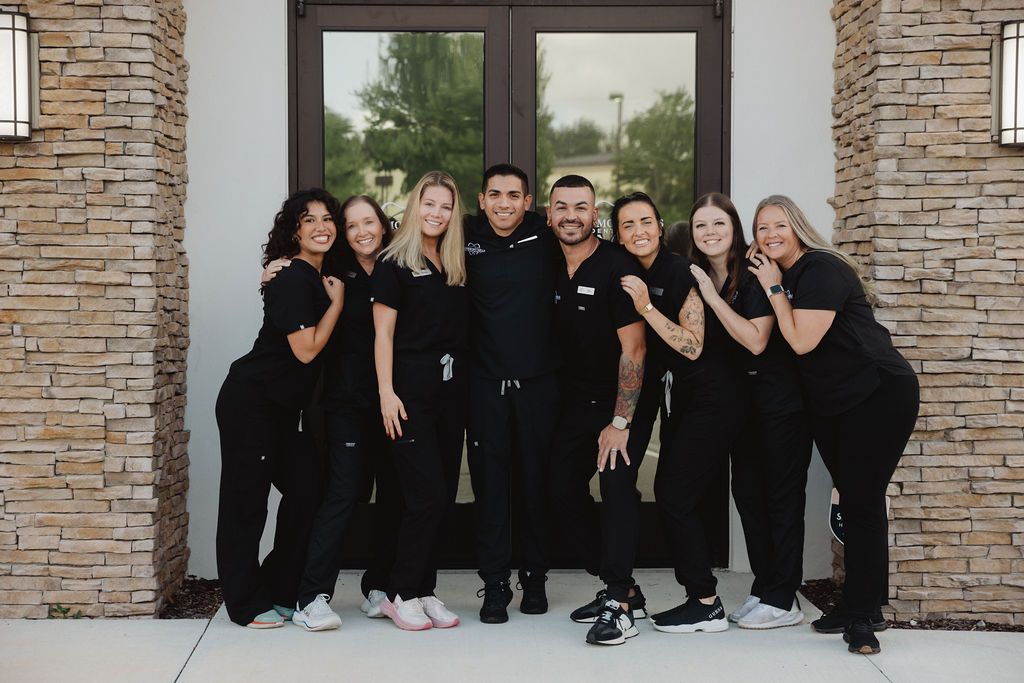 Group of dental office staff posing in front of a building with stone columns. They wear black scrubs and sneakers.