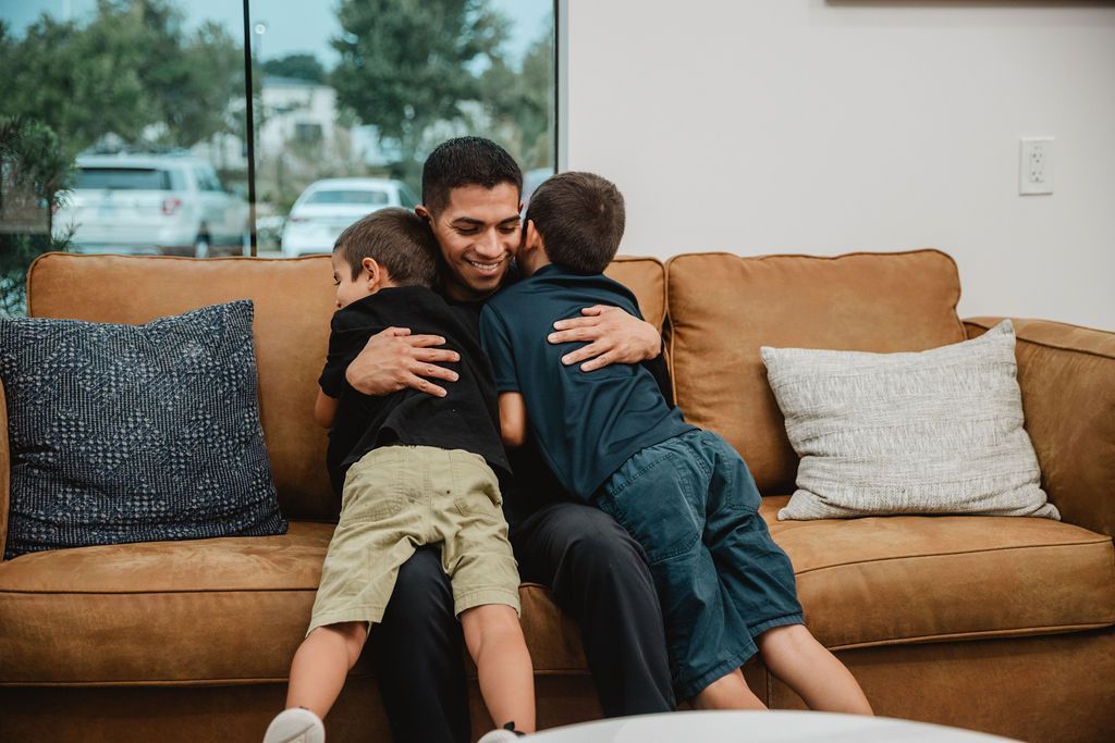 Man sitting on a brown couch hugs two children indoors near a window.