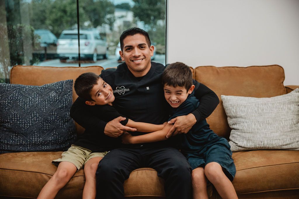 Man smiles, embracing two boys on a brown sofa. Interior setting.