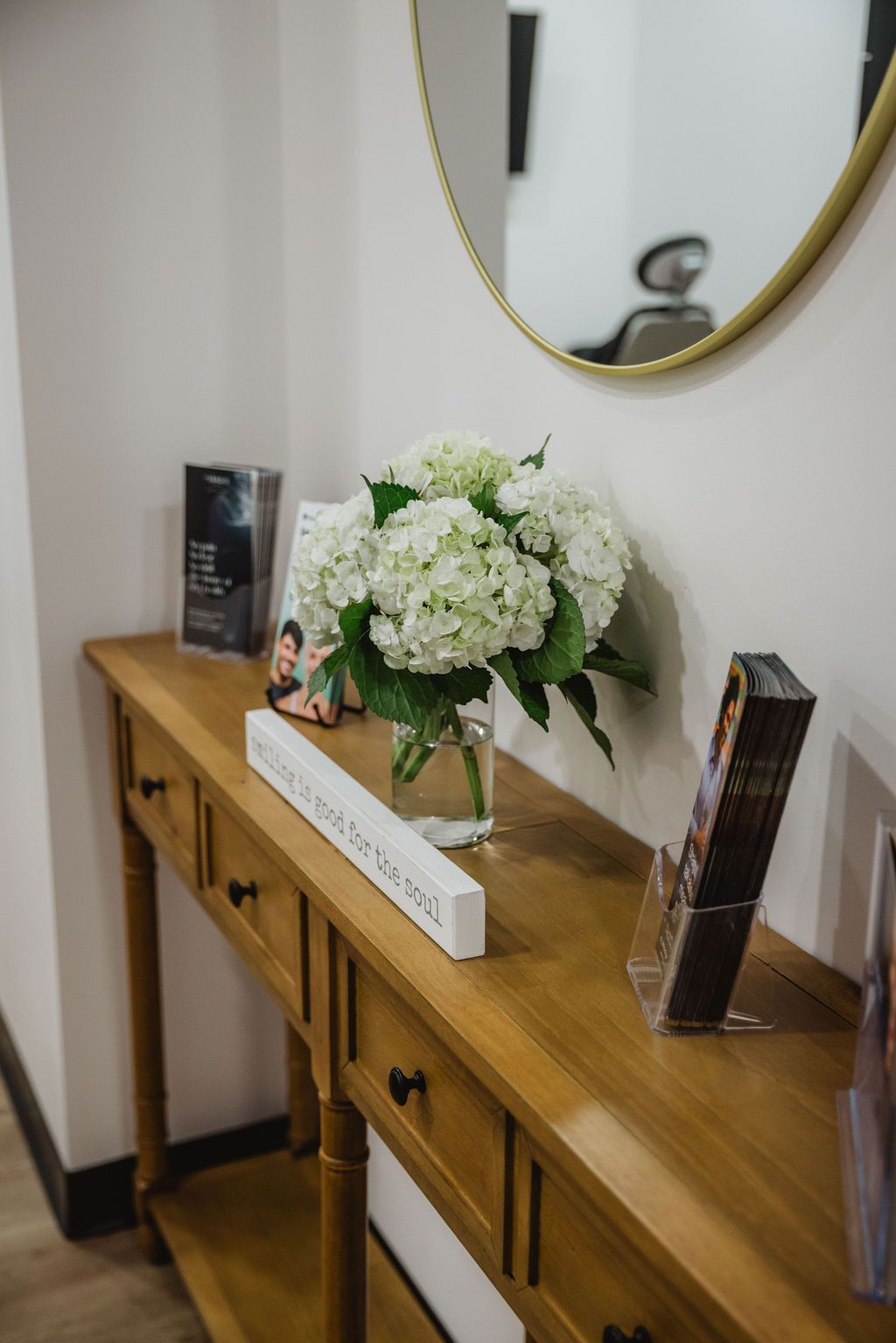 Wooden console table with white hydrangeas, mirror, and brochures in a bright room.