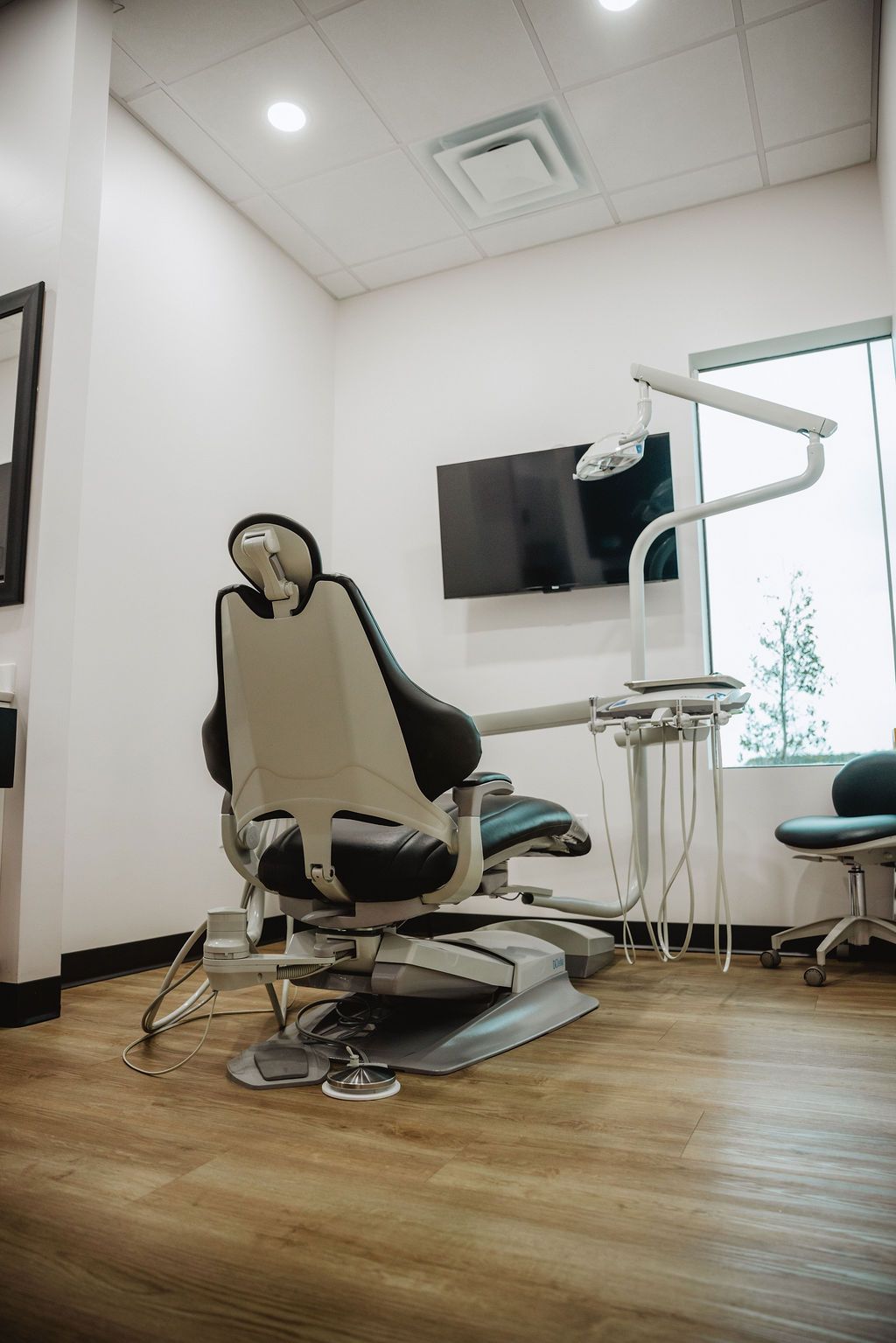 Dental examination room: dental chair, mounted monitor, equipment, large window, wood flooring.