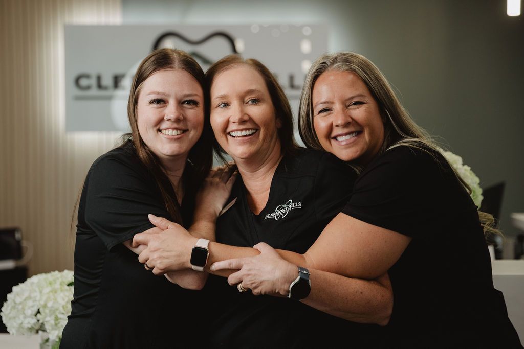 Three smiling women in black shirts embrace, in a dental office setting.