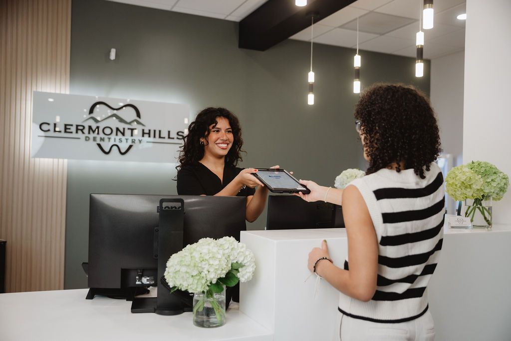 Receptionist at a dental office hands a tablet to a patient. Brightly lit, white desk, smiling.