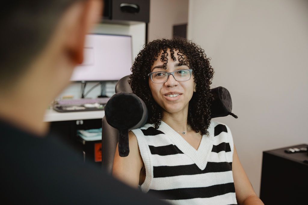 Woman in glasses smiles, sitting in chair. Striped vest, indoor setting, talking to a person out of view.