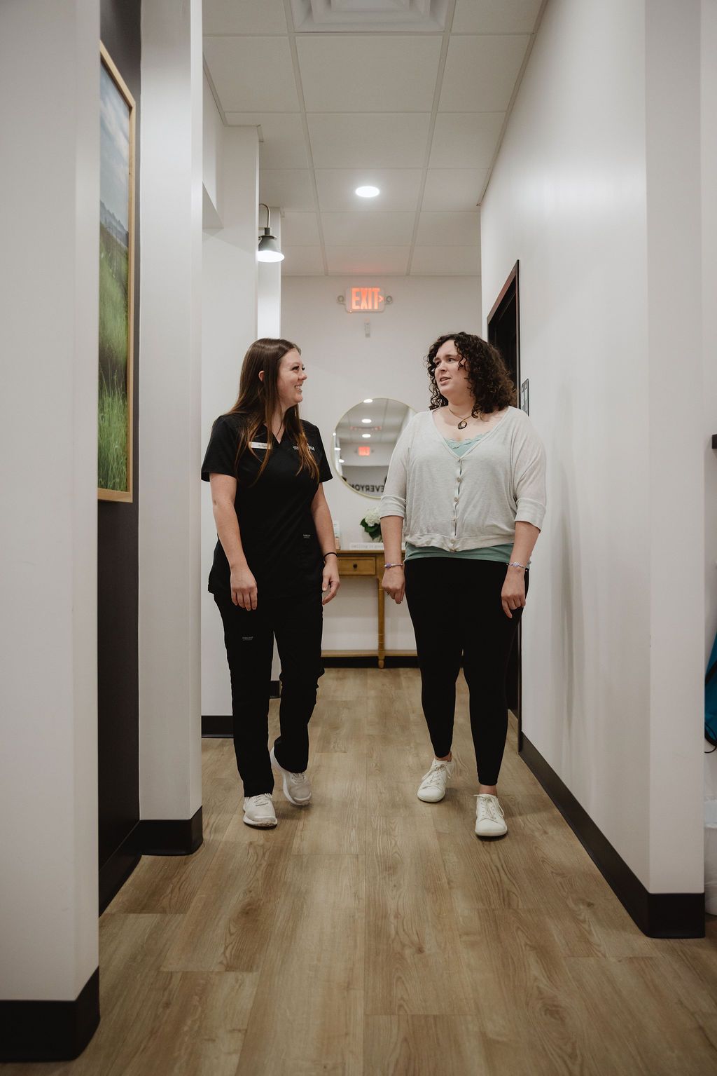 Two people walking in a hallway, one in scrubs, smiling; other person in casual clothes, smiling.