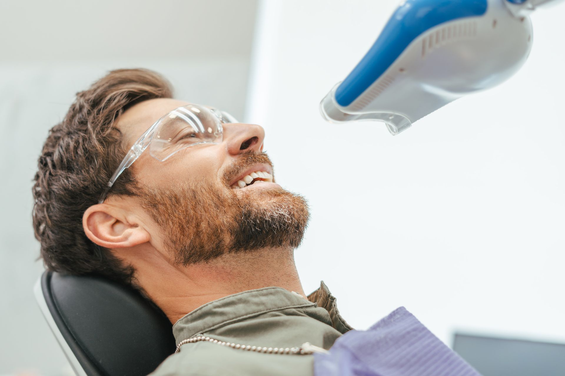 Man in dentist chair with mouth open, receiving teeth whitening treatment under a blue light.