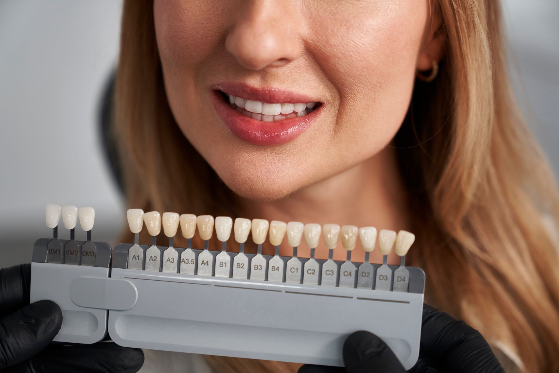 Woman holding a dental shade guide to match tooth color; smiling.