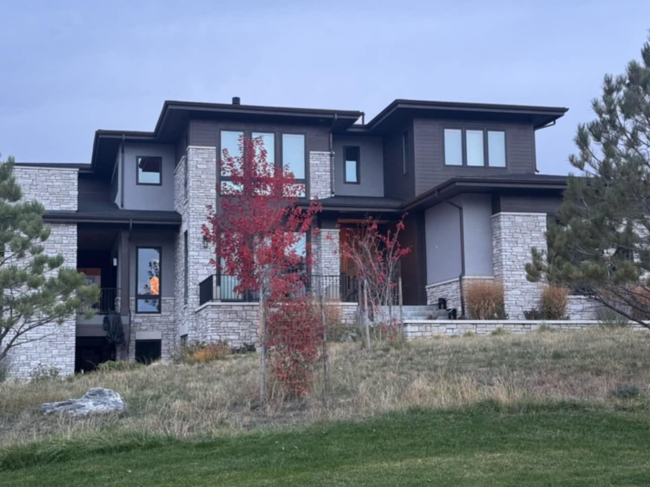 Modern two-story house with gray and stone exterior, large windows, and a red tree in front.