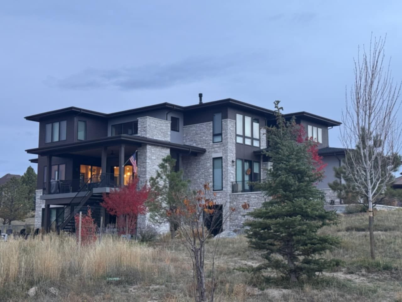 Two-story stone home with dark trim and large windows, surrounded by trees and dry grass under a cloudy sky.