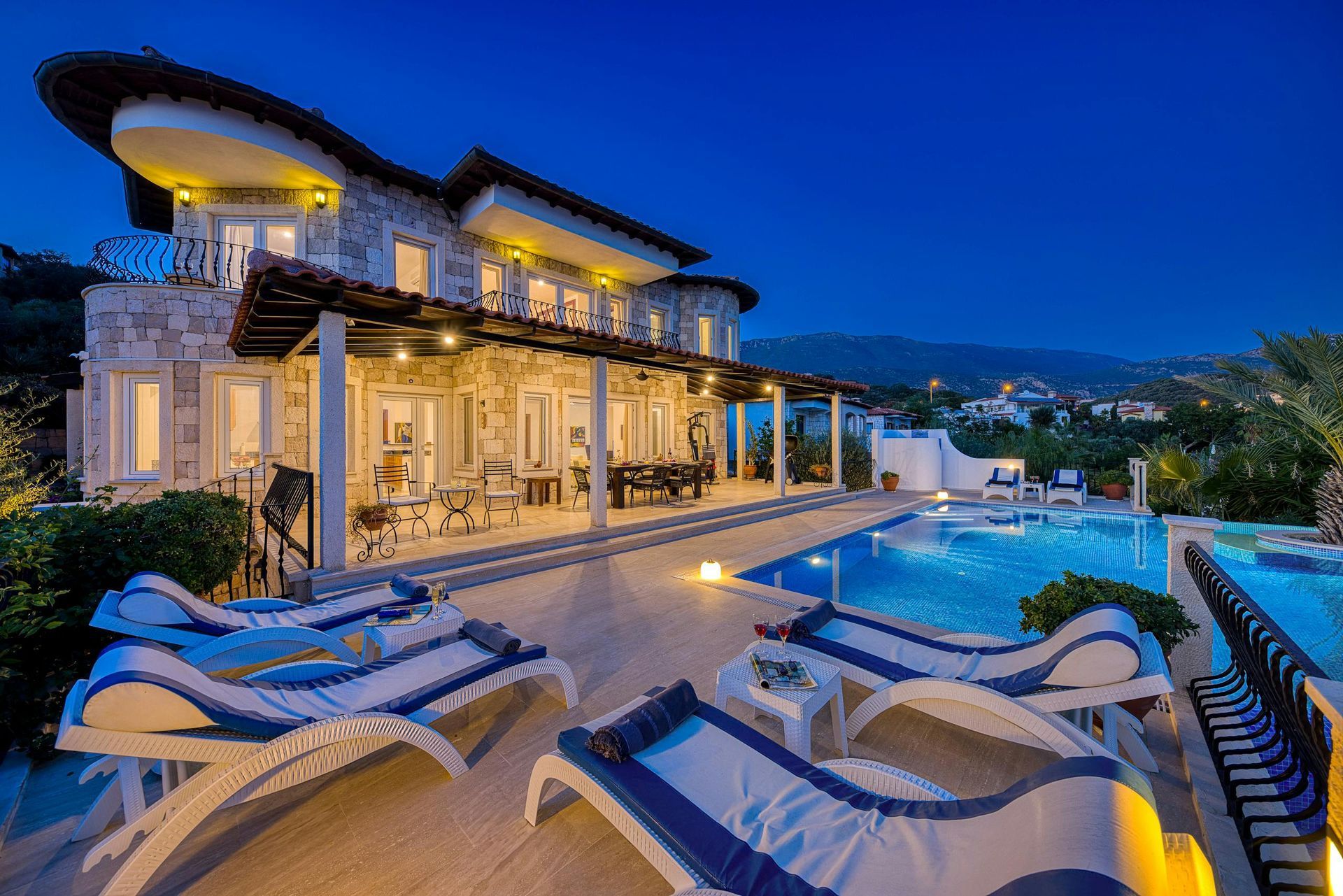 Luxury villa with pool at dusk, lit up; lounge chairs, mountain view.