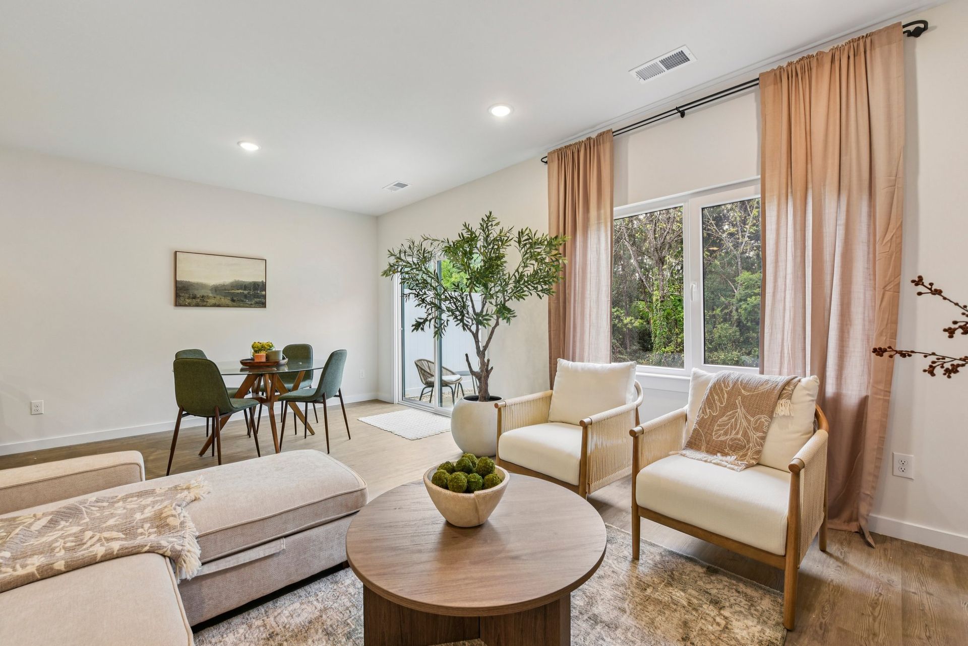 Living room with dining area. White walls, light wood floors, neutral furniture, large window with pink curtains.