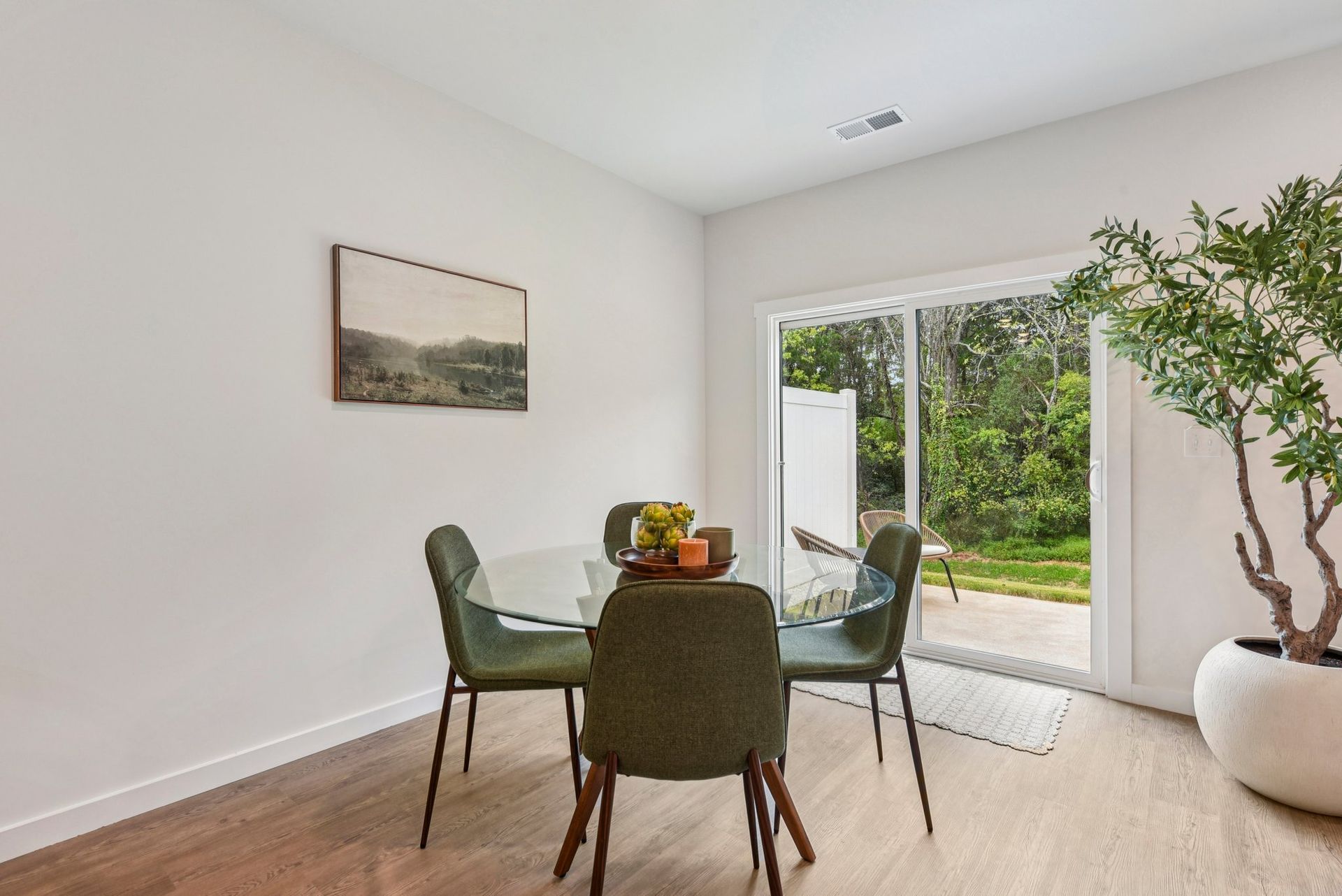 Dining room with glass table, green chairs, art, sliding door to backyard, and potted tree.