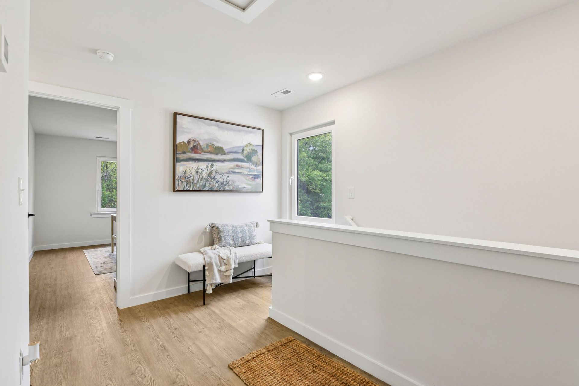 Hallway with wooden floor, white walls, bench, artwork, and window overlooking trees.