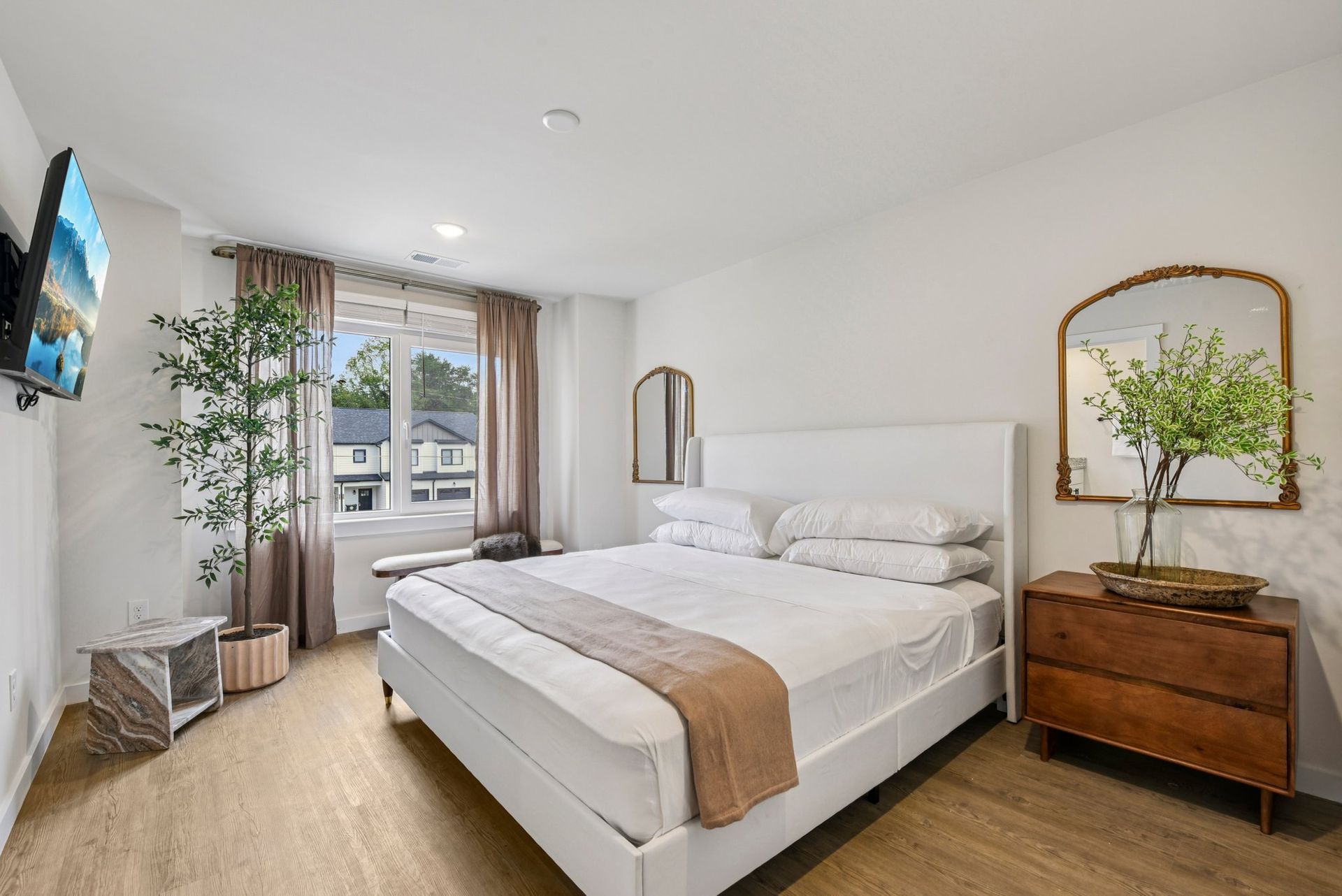 Bedroom with white bed, wooden dresser, large mirror, and TV on wall.