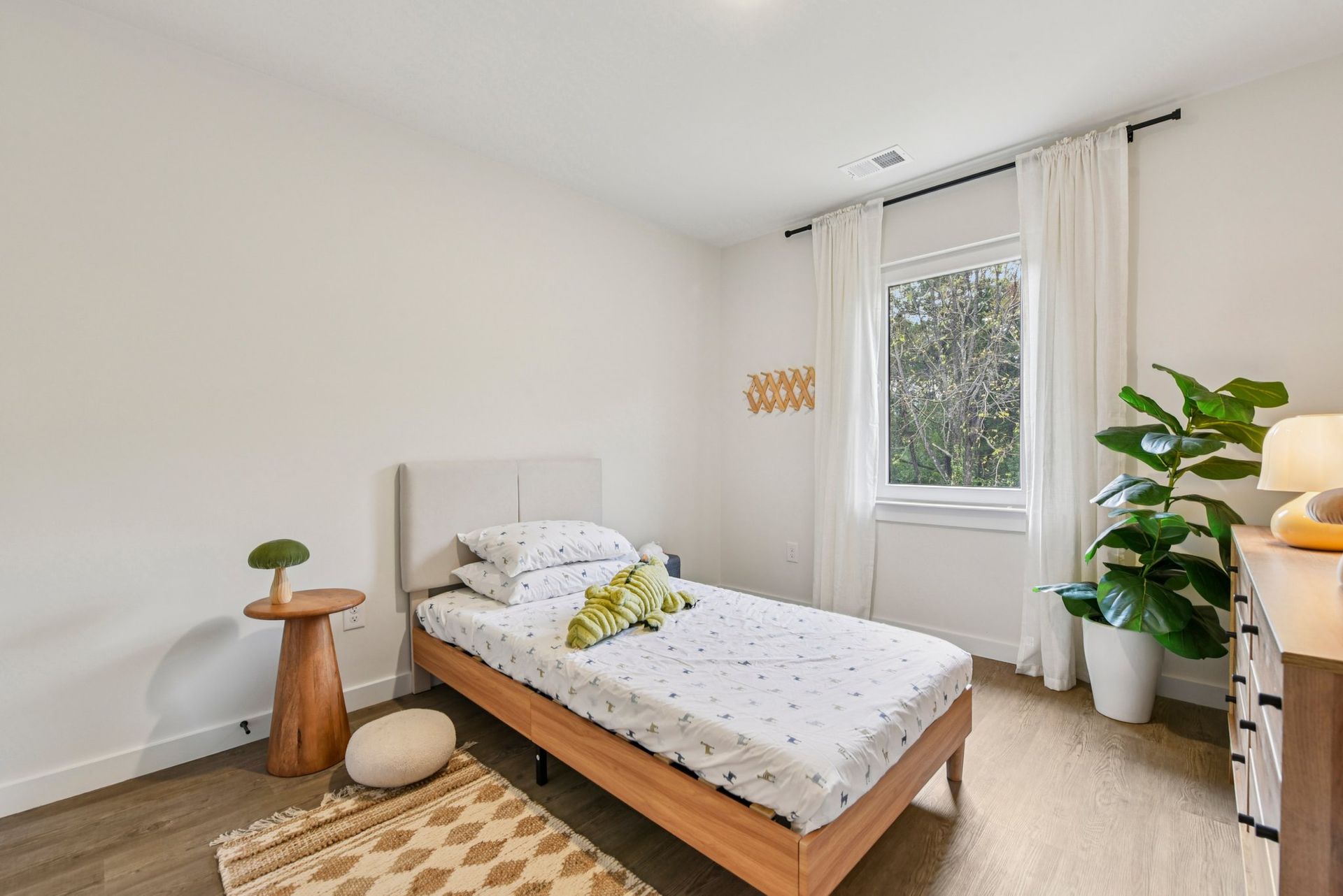 Bedroom with white walls, a single bed, a window with curtains, and a side table with a plant.