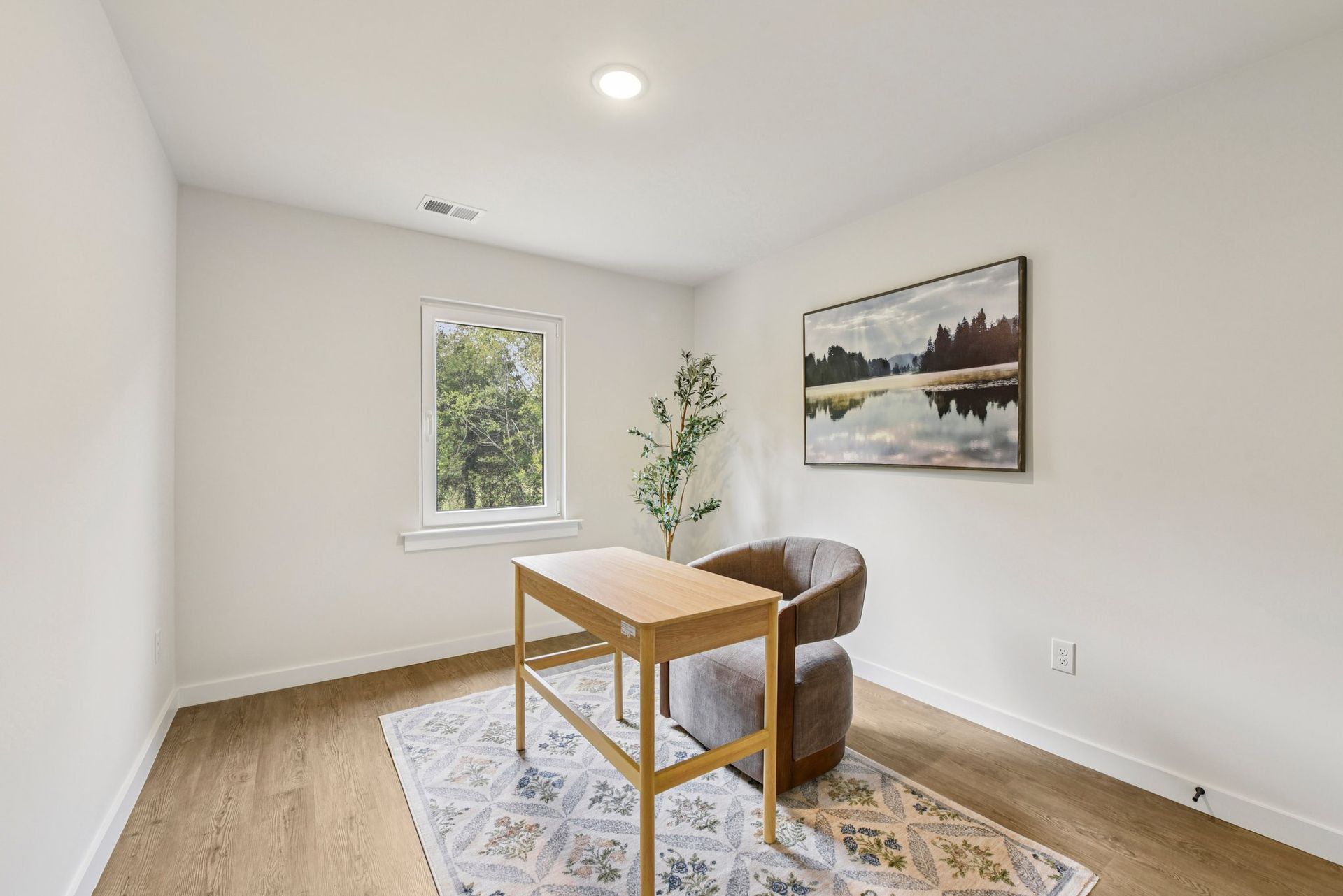 Home office with wooden desk, chair, and art, light wood floors, window.