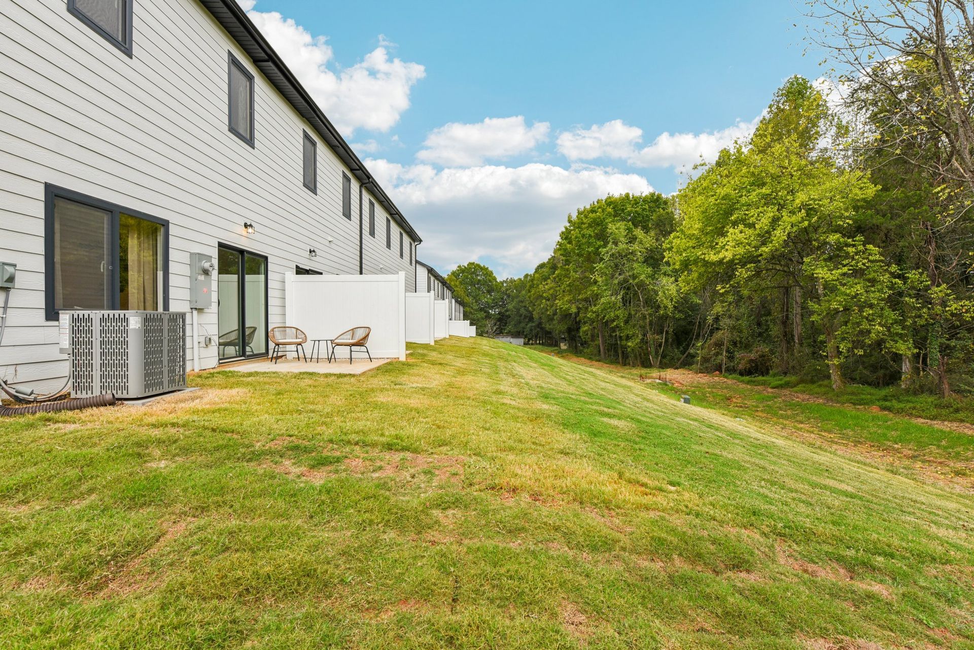 Row of white townhouses with patios, next to a grassy hill and trees under a blue sky.