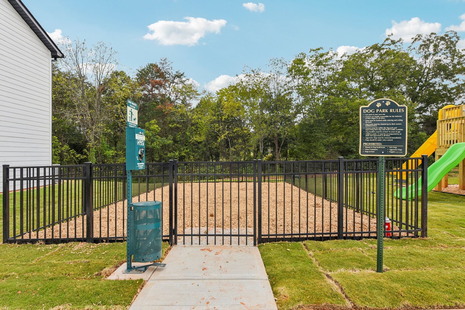 Dog park with black fence, mulch, waste bag dispenser, and informational sign; green trees in the background.