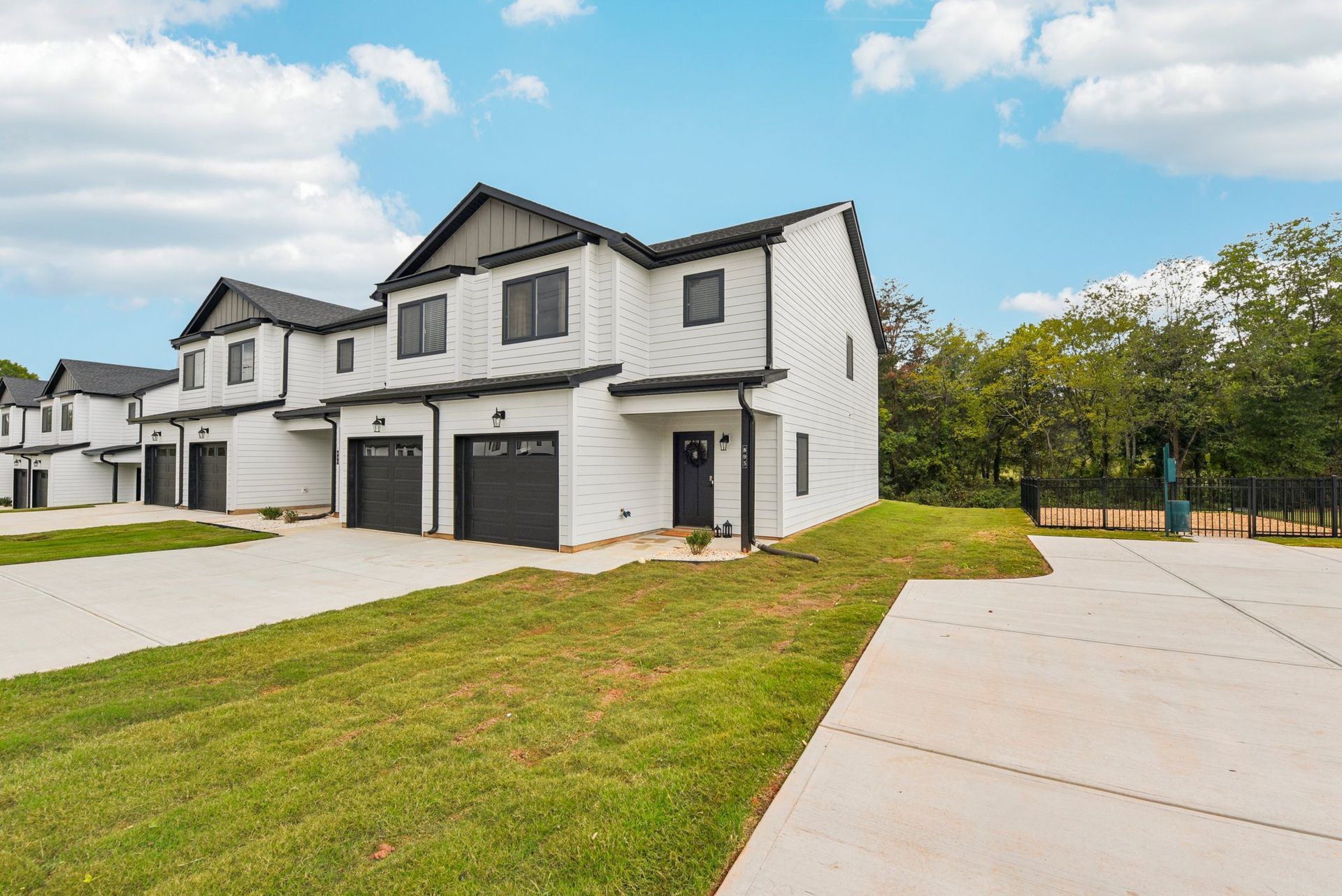 White townhomes with black accents, garages, and driveways, set on a green lawn under a blue sky.