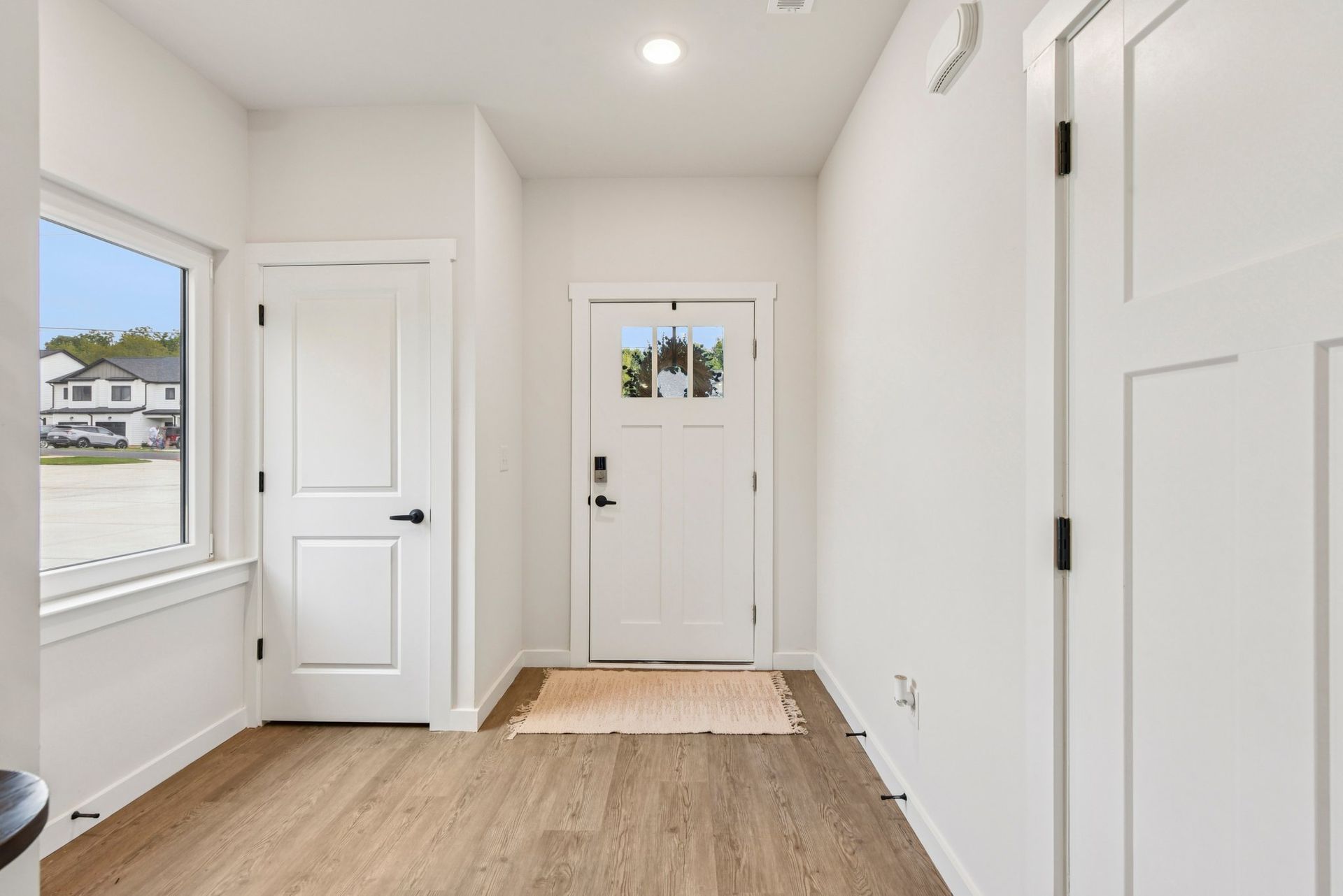 Bright white entryway with front door, doors to rooms, window. Beige floor mat.