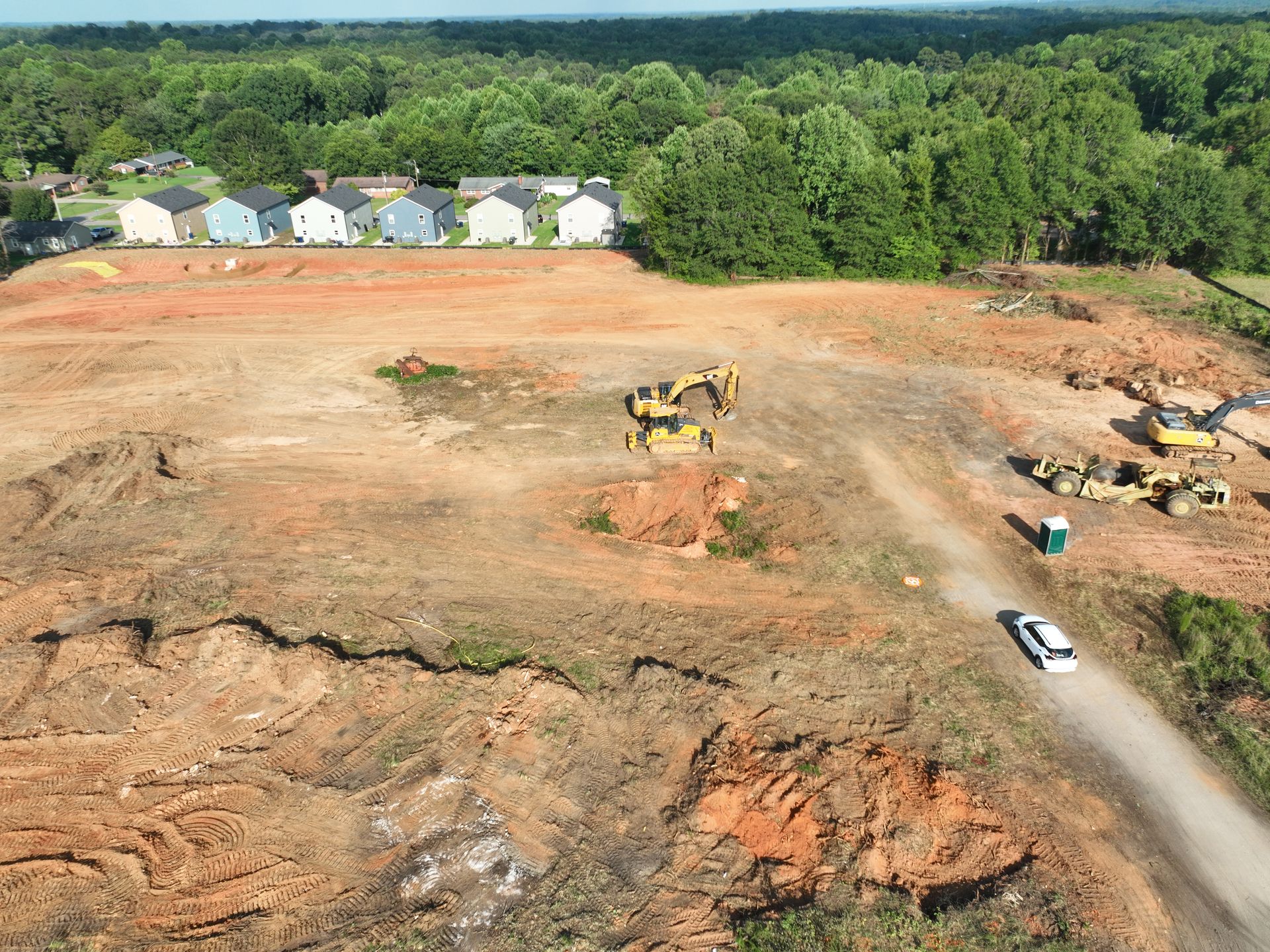 Construction site with heavy machinery, cleared red soil, and a row of houses in the background.