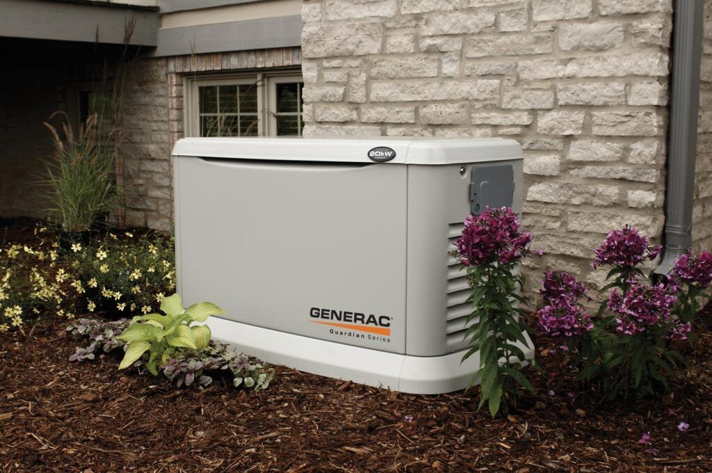Gray Generac home backup generator near a brick house with flowers.