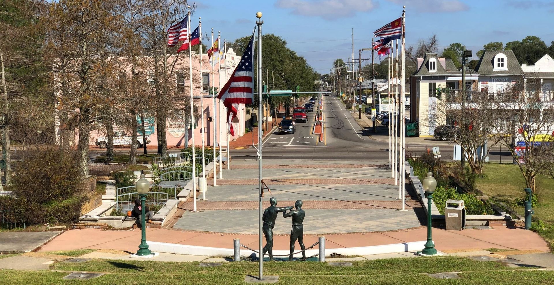 Street view with flags, statues, and buildings in a town square, vehicles on the road. Kenner