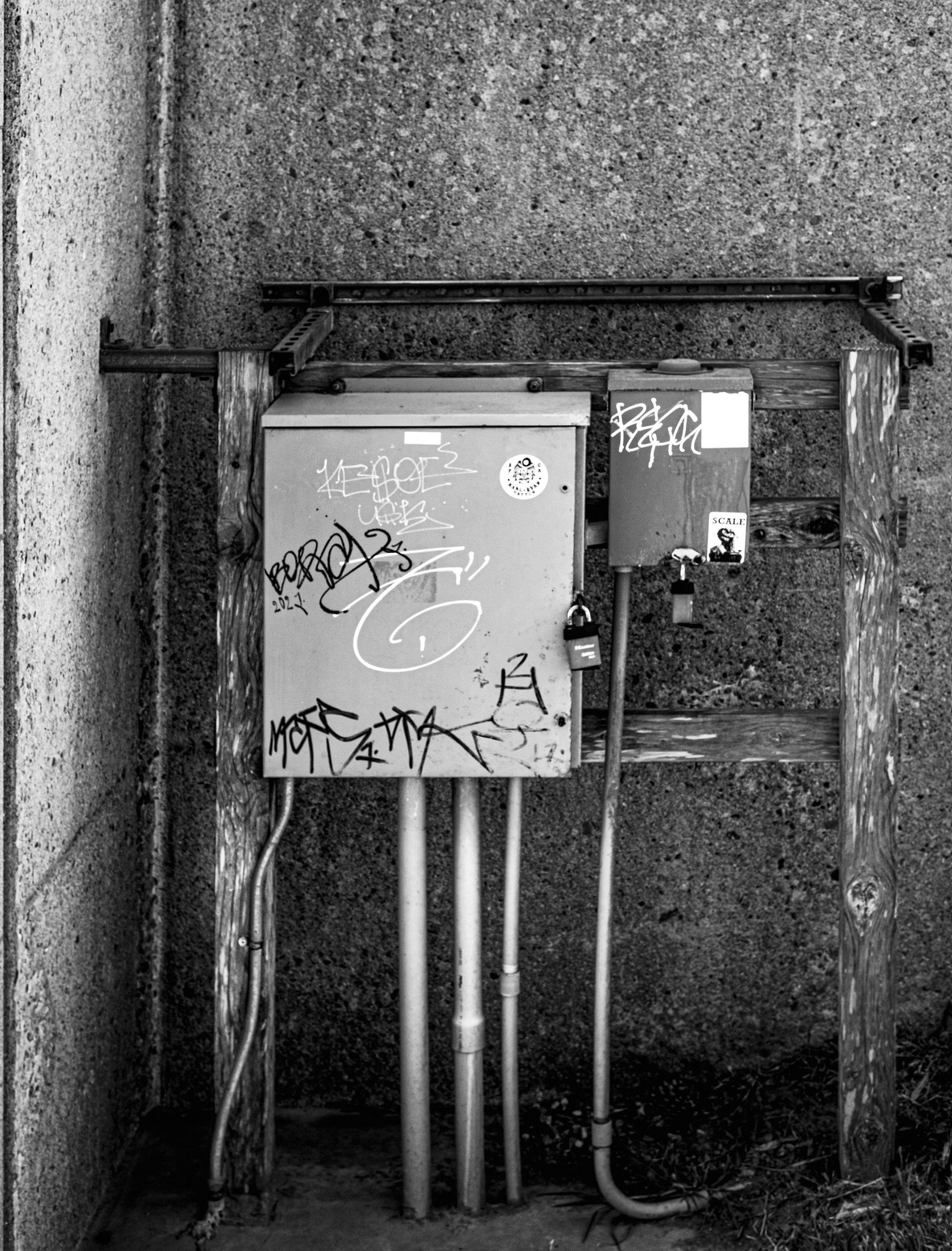 Black and white photo of a graffitied electrical box and conduit pipes against a textured wall.