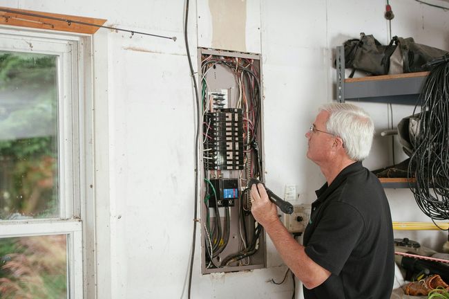 Man inspects electrical panel in a garage, using a tool. The panel's wiring is visible, and the setting is bright.