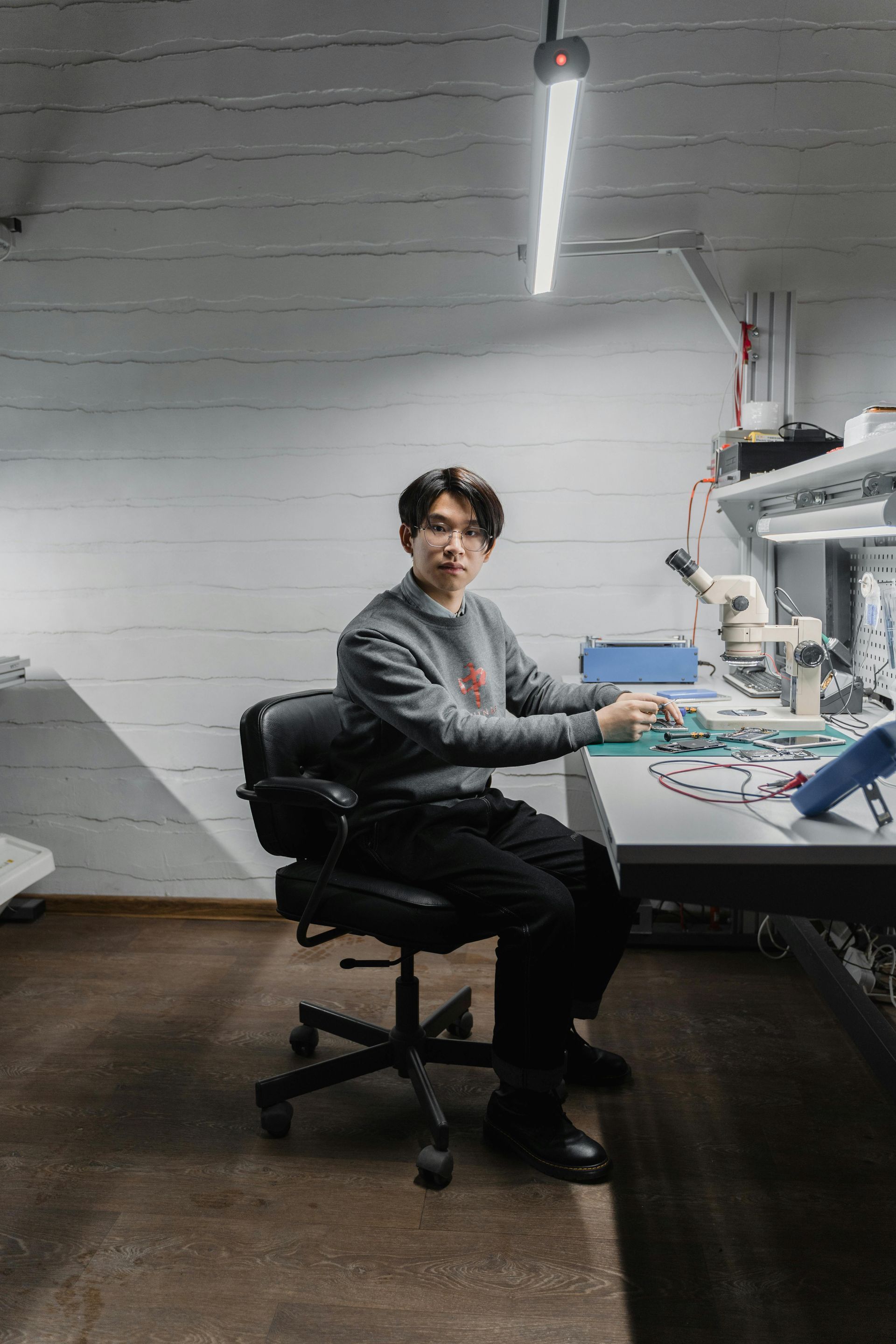 Man seated at a workbench, looking at the viewer. He wears a gray sweater and is in a workshop with electronics.