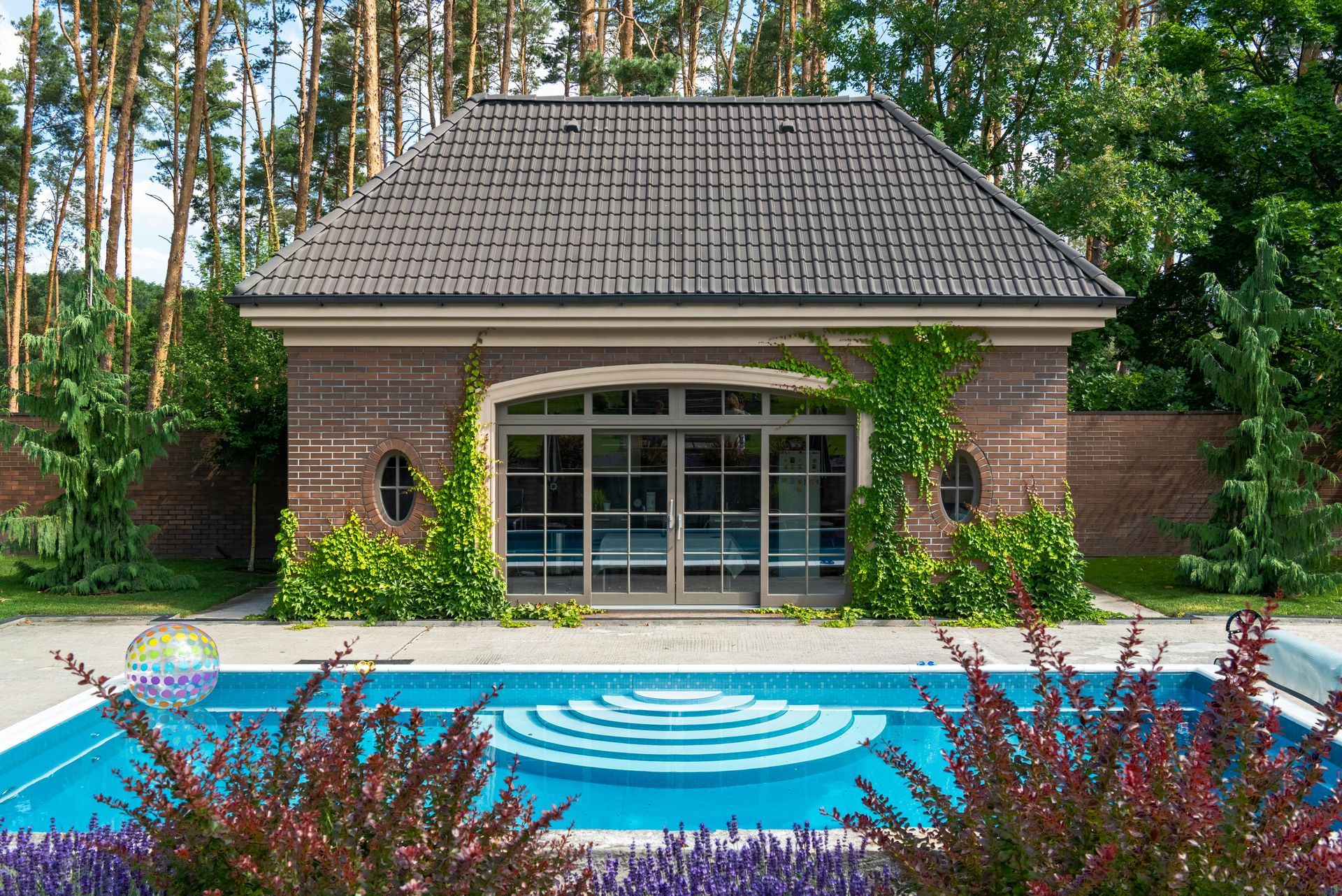 Poolside brick building with glass doors, climbing vines, and blue pool with steps.