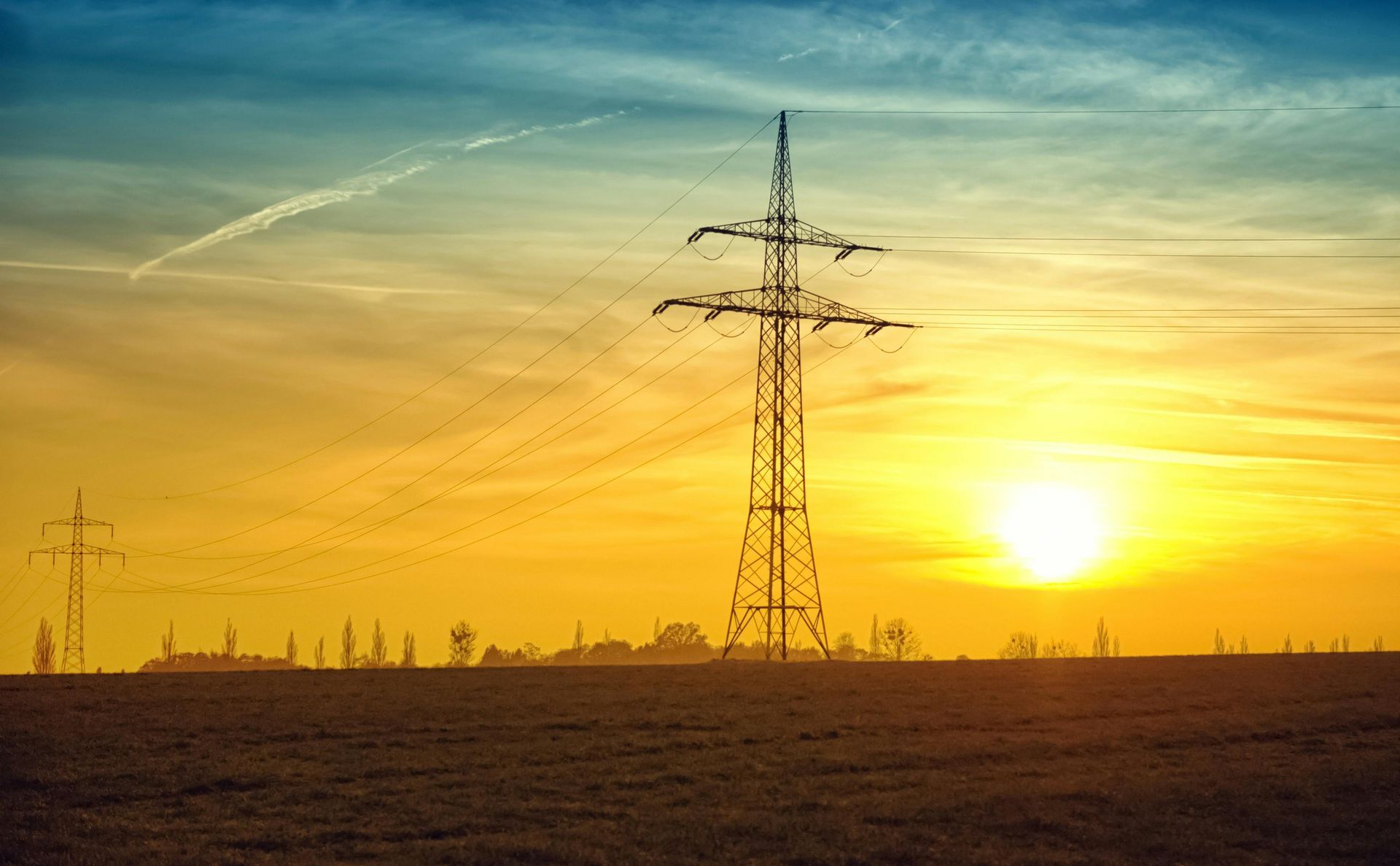 Power lines silhouetted against a vibrant sunset over a field; the sun is orange and yellow, with some clouds.