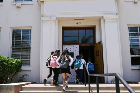 Students entering a white building through a front doorway, carrying backpacks and bags.