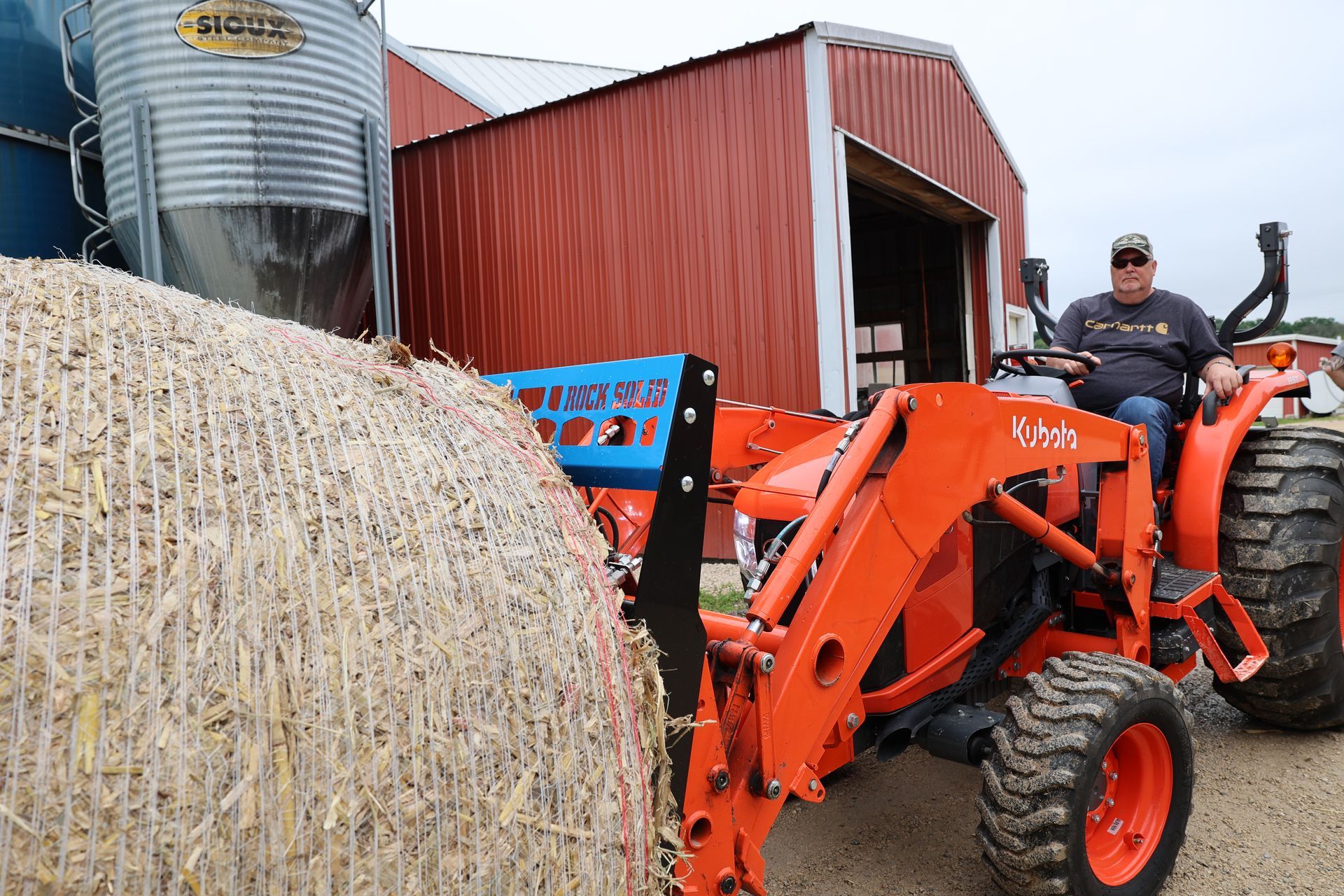 A man is driving an orange tractor next to a bale of hay.