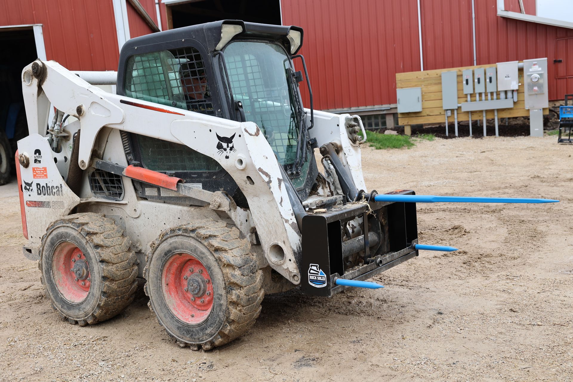 A bobcat tractor is parked in a dirt field in front of a red barn.