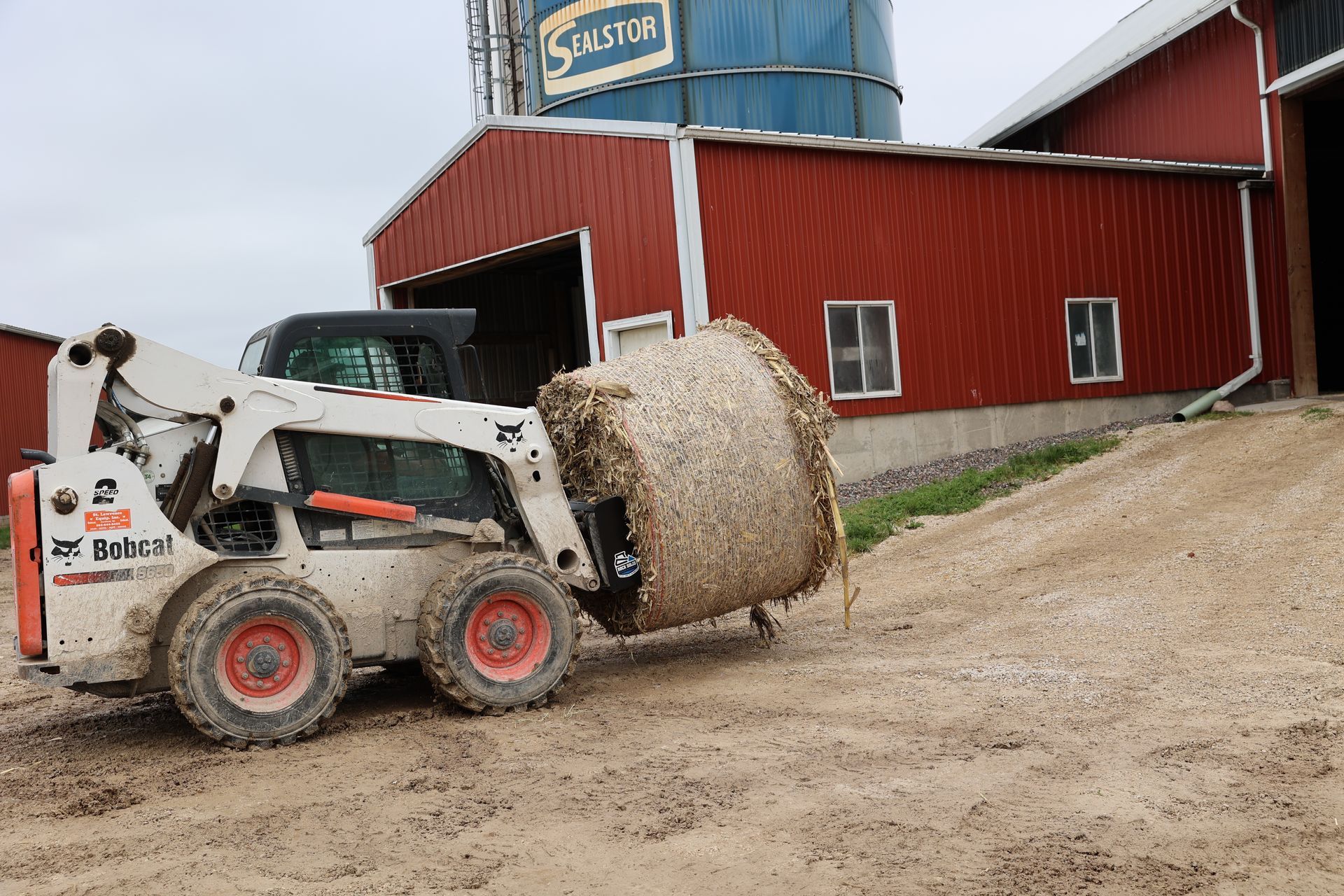 A bobcat is carrying a bale of hay in front of a barn.