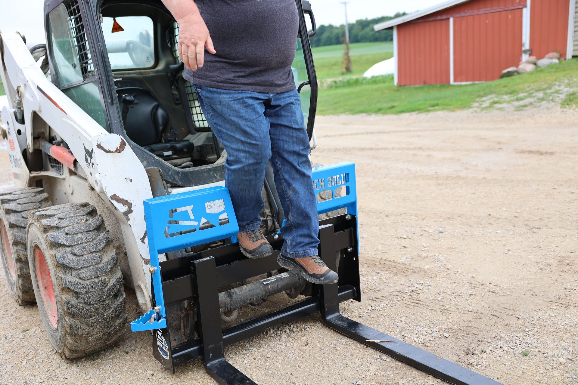A man is standing on a skid steer with forks attached to it.