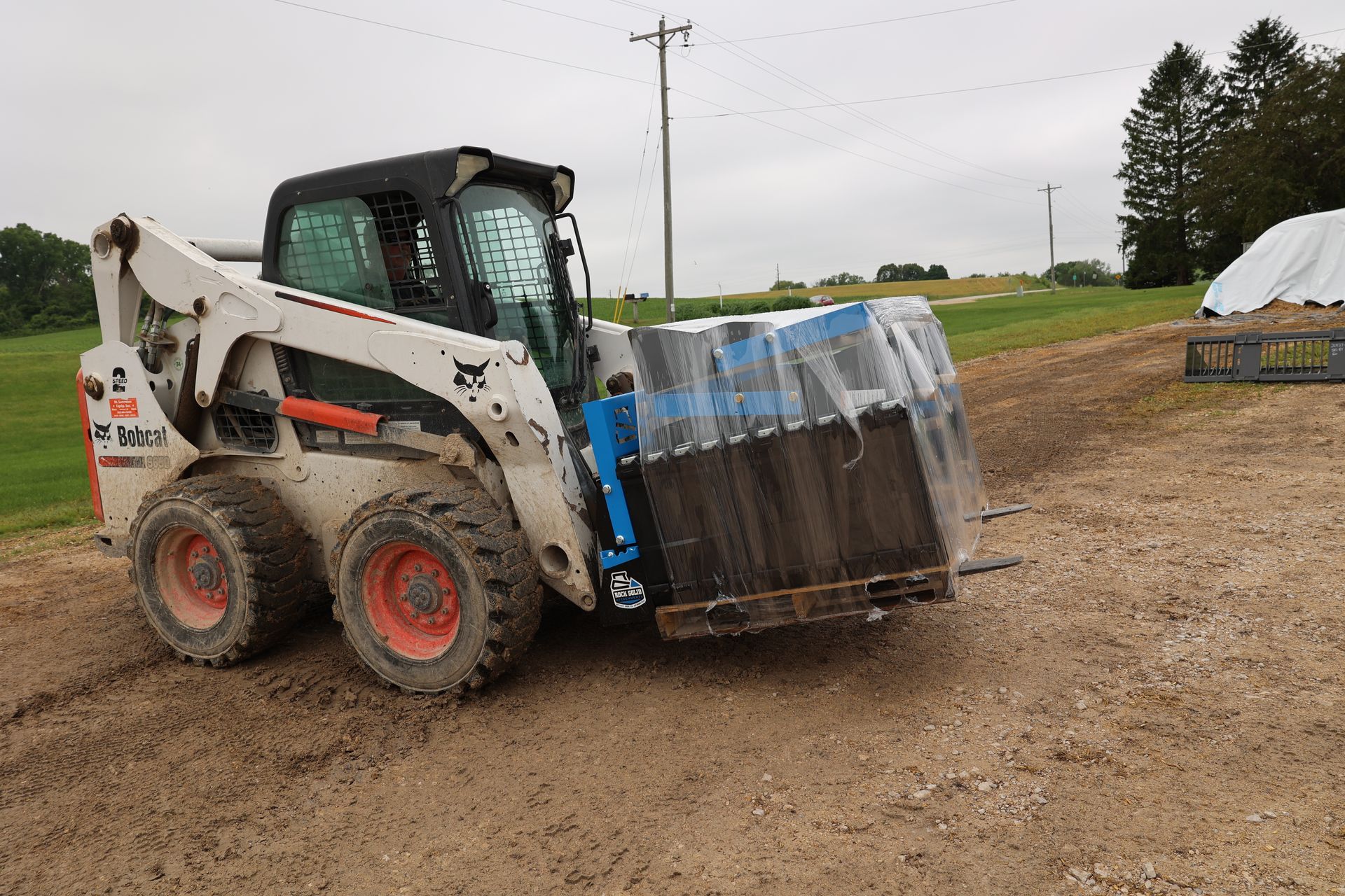A bobcat is carrying a pallet of bricks on a dirt road.