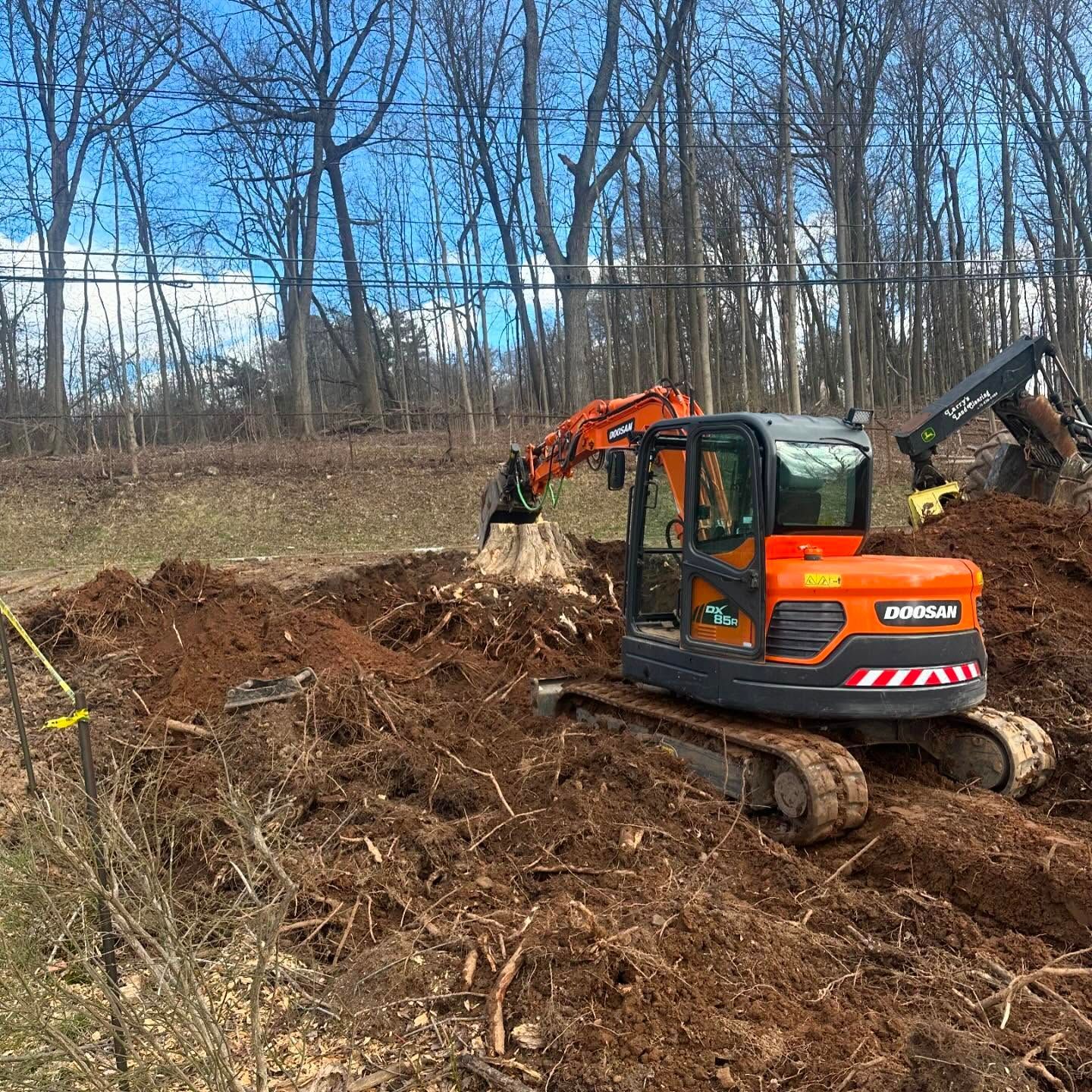 Orange excavator digging in a dirt pile, with another excavator in the background, blue sky.